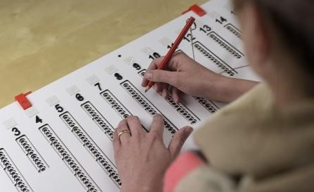 Een plaatje van een mal die over het stembiljet gelegd wordt, met de handen van een slechtziende die het rode potlood vasthoudt.