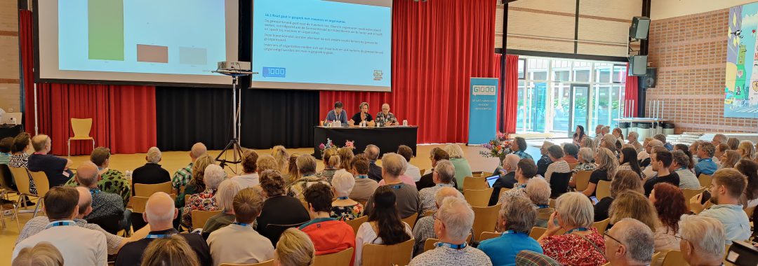 Zaal met mensen en twee grote schermen voor de Burgerraad in Schagen